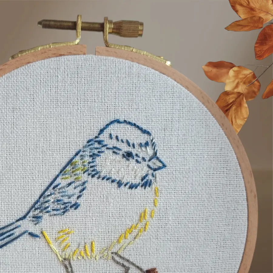 Motif brodé à la main d’une mésange aux plumes bleues et jaunes, délicatement posée sur une branche, présenté dans un tambour à broder en bois..