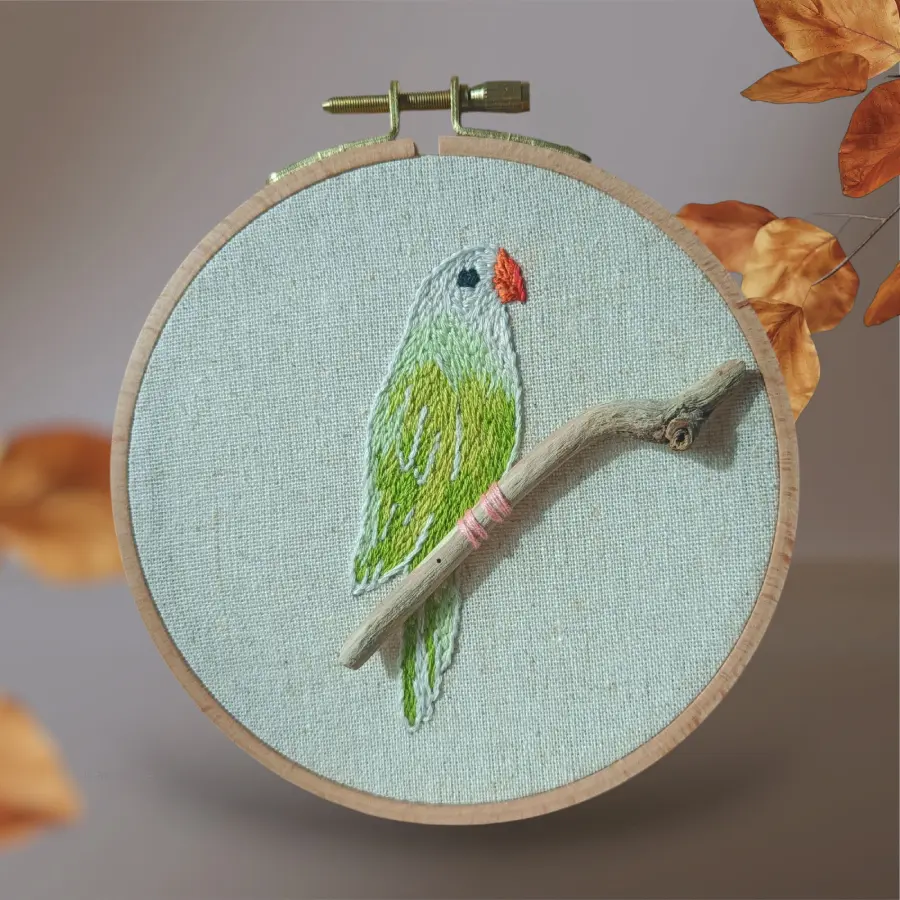 Broderie d’une perruche verte au bec rouge, perchée sur une petite branche en bois, présentée dans un tambour à broder rond.