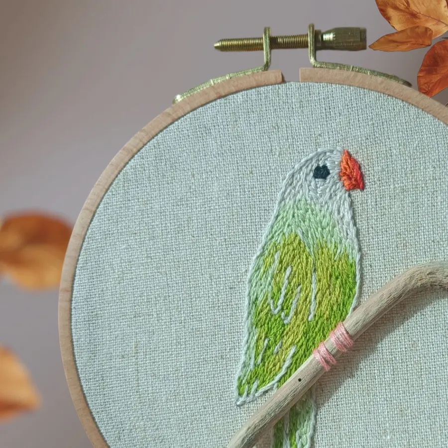 Broderie d’une perruche verte au bec orangé, perchée sur une petite branche en bois